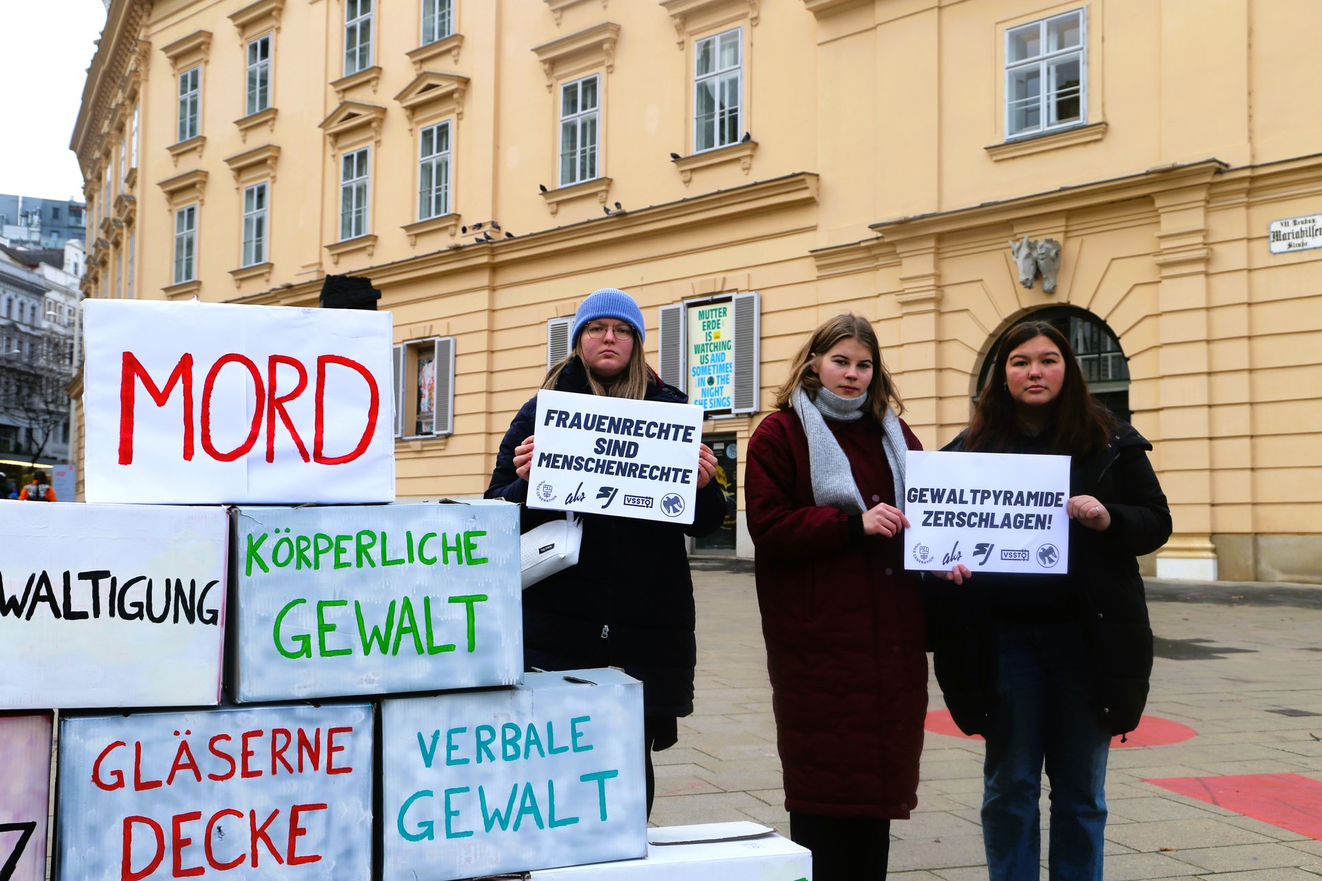 Die Gewaltpyramide wird mit Kartons dargestellt. Auf der linken Seite der Pyramide stehen drei Frauen und halten Schilder. Auf den Schildern steht: "Frauenrechte sind Menschenrechte" und "Gewaltpyramide zerschlagen".