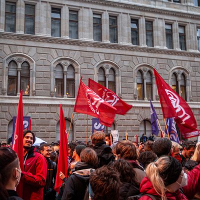 Demonstration. Die Straße ist voll mit Menschen, mehrere Fahnen sind zu sehen.
