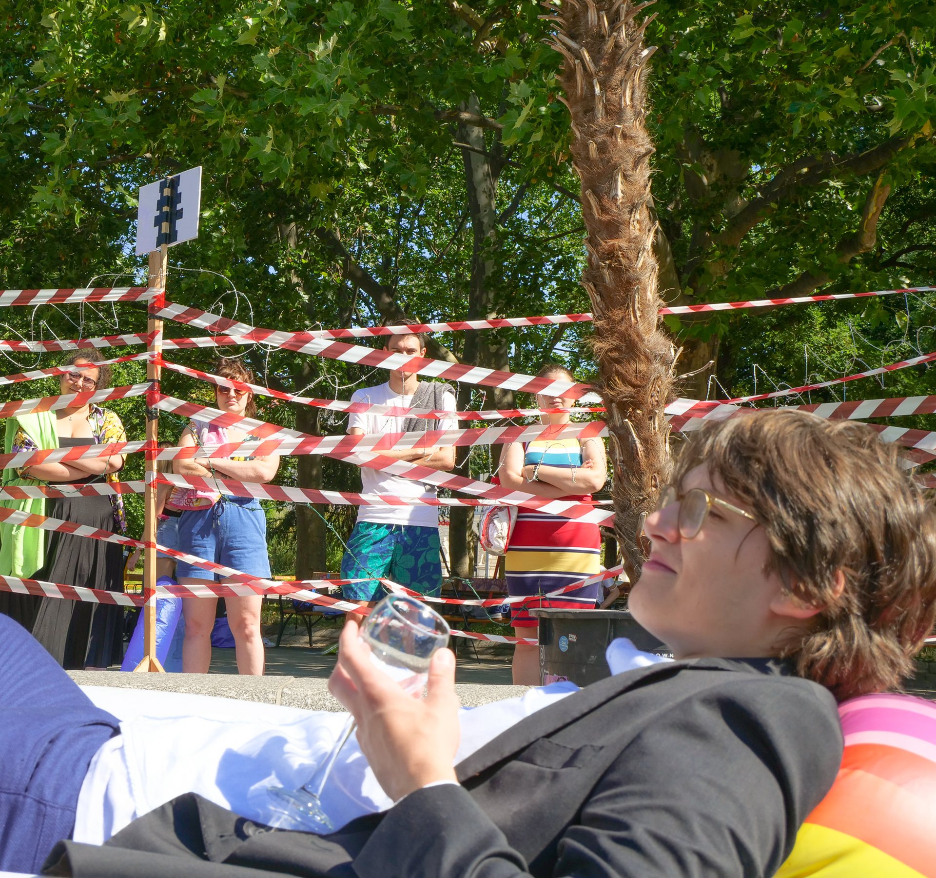 Ein Mann liegt auf einer Luftmatratze und genießt die Sonne. Vier andere Jugendliche stehen vor einem Absperrband und sind vom Badeplatz ausgesperrt.
